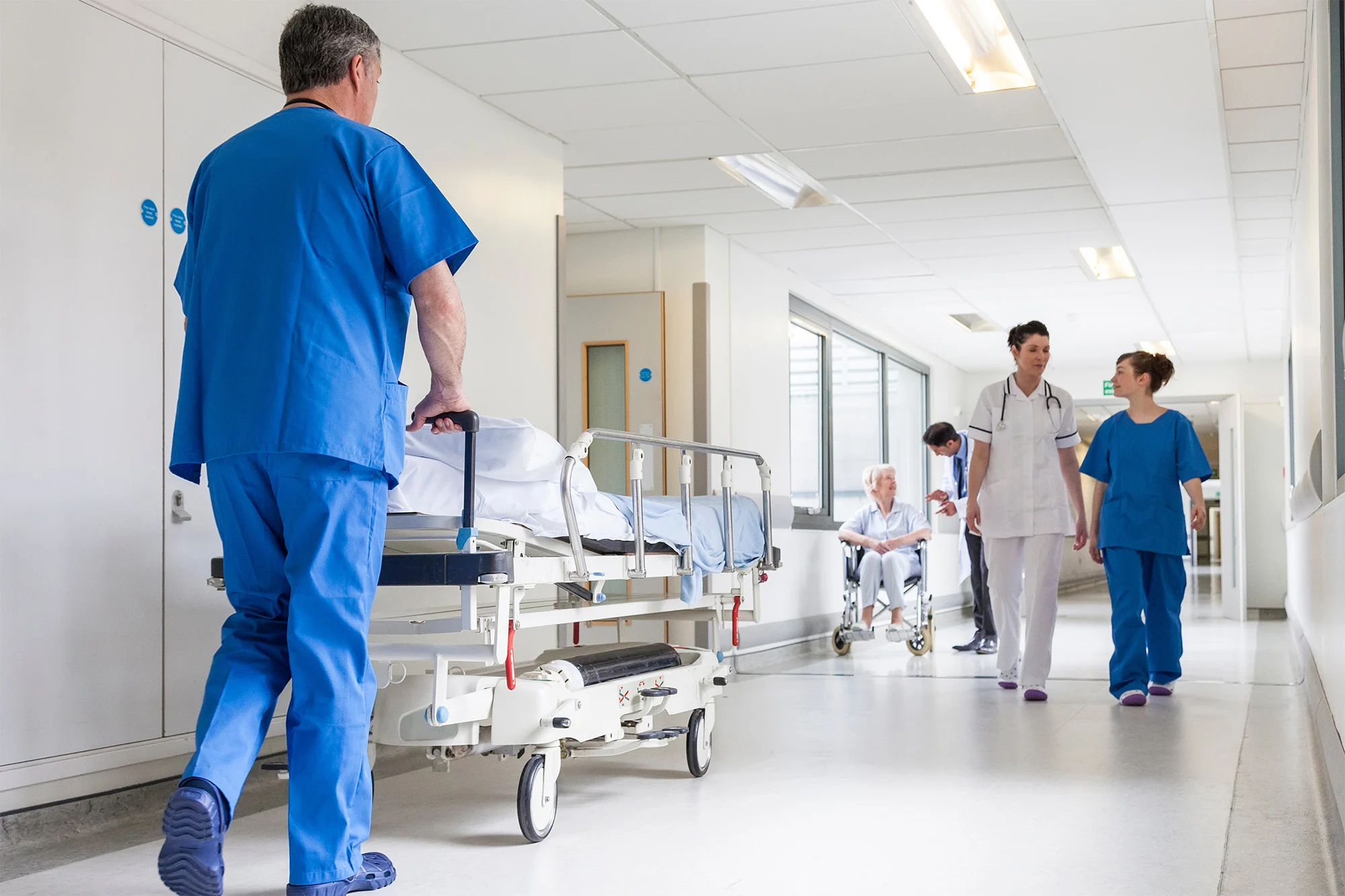 Pflegekraft schiebt Krankenhausbett durch hellen Klinikflur, im Hintergrund medizinisches Personal im Gespräch.