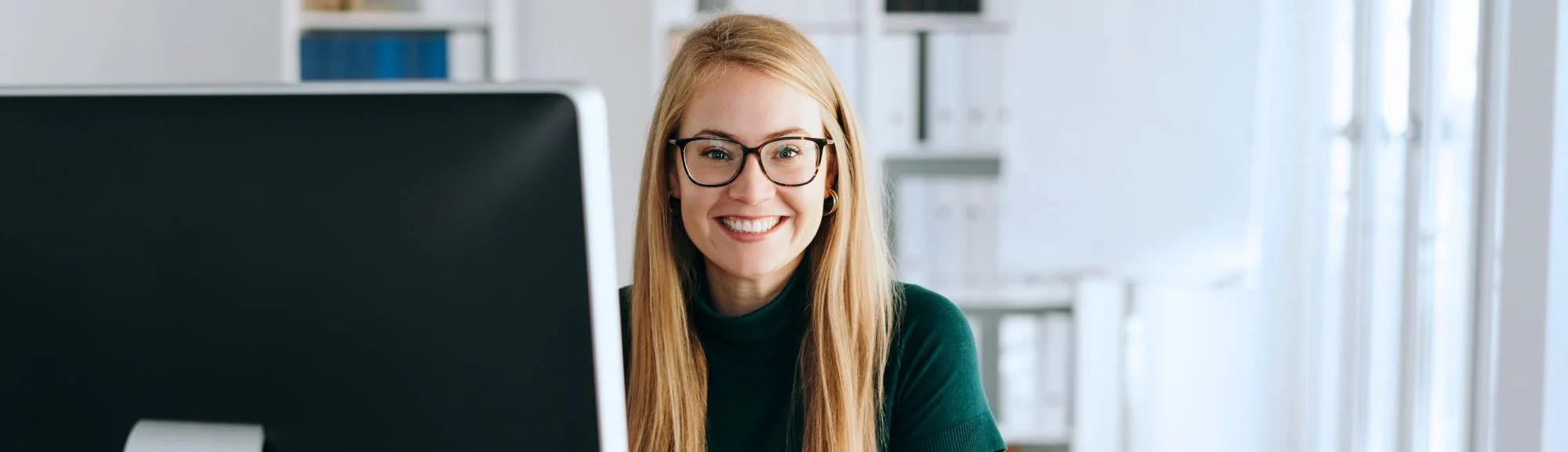 Junge Frau mit langen blonden Haaren und schwarzer Brille sitzt lächelnd an einem Schreibtisch vor einem Computermonitor in einem hellen Büro.