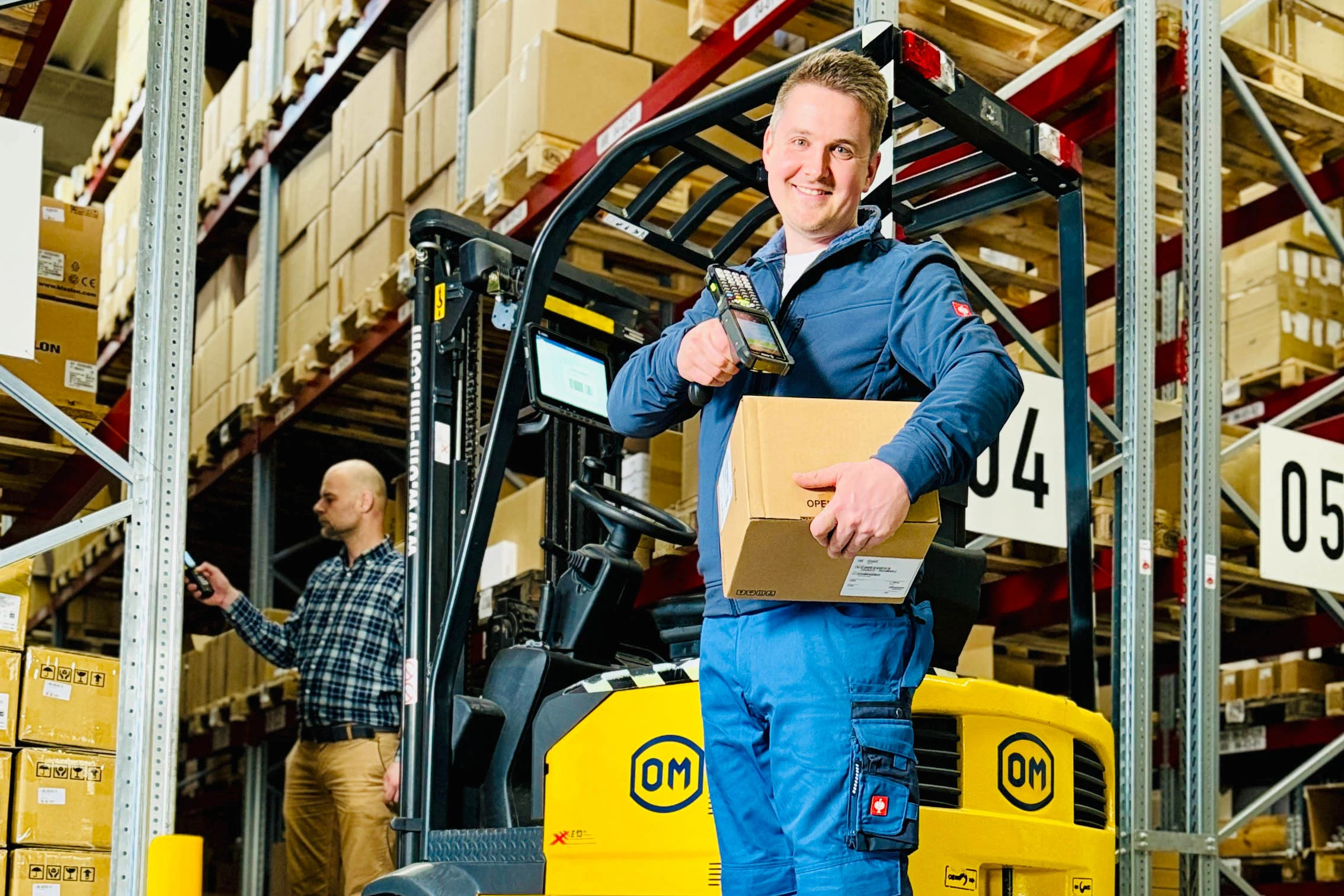 Lagerarbeiter in blauer Arbeitskleidung steht mit einem Paket und einem Barcodescanner vor einem gelben Gabelstapler in einem Hochregallager, während im Hintergrund ein Kollege Inventur macht.